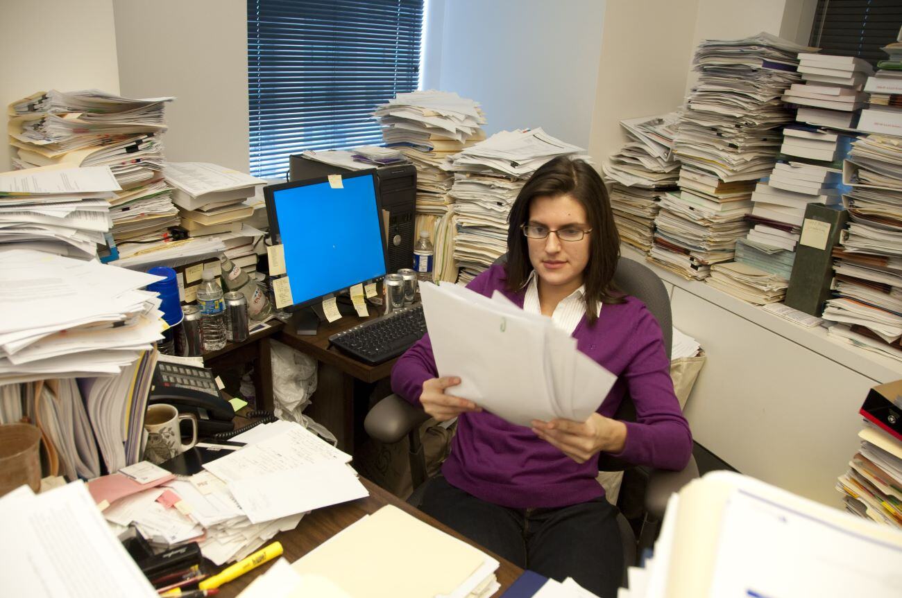 woman sits at a cluttered desk in an office, surrounded by tall stacks of papers and files